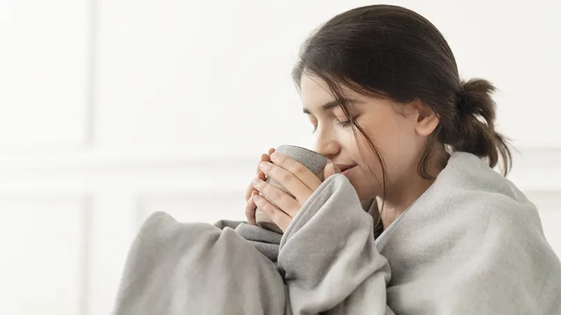 Image d'une femme buvant du thé enveloppée dans une couverture lestée pour une stimulation par pression profonde.