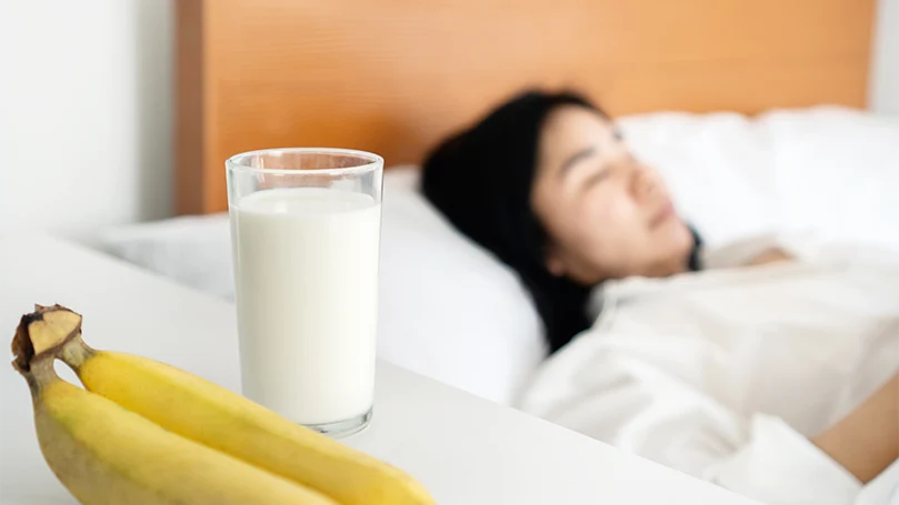 L'image d'une femme qui dort à côté d'une table de nuit sur laquelle se trouvent du lait et des bananes.