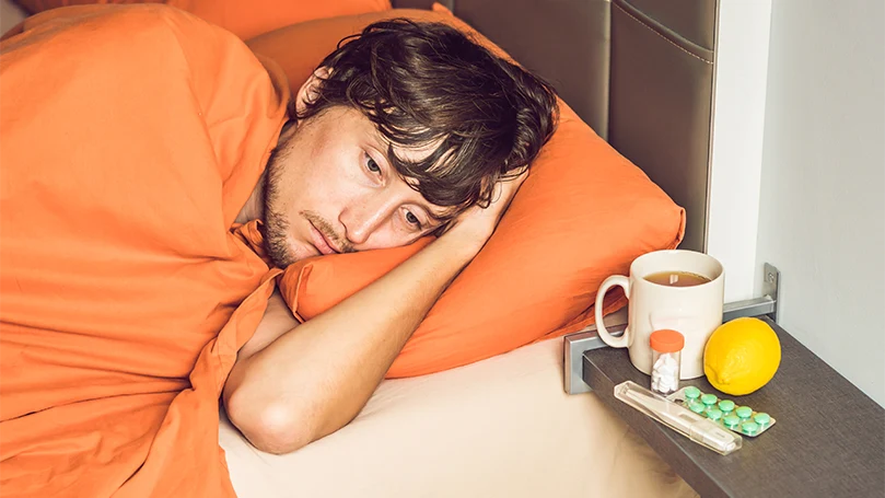 Un homme allongé dans un lit malade et recouvert d'une couverture orange.