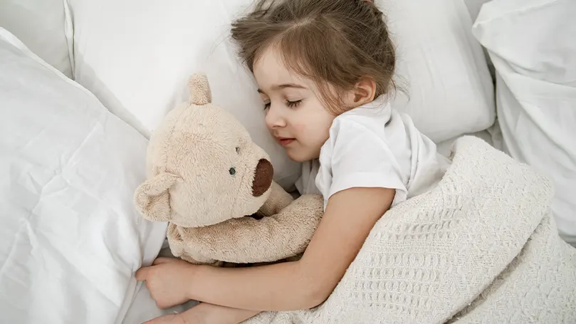 Image d'une petite fille confortablement installée dans son lit avec son animal en peluche.