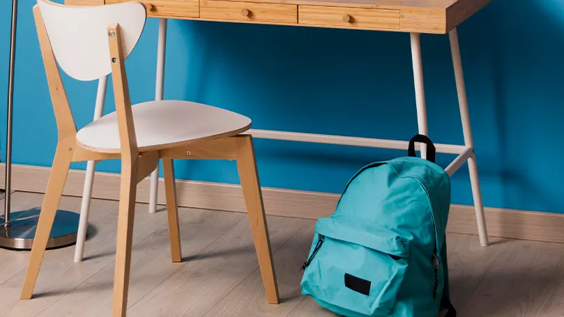 Mur bleu, sol en bois avec un bureau et une chaise en bois. Un sac à dos bleu est placé devant.